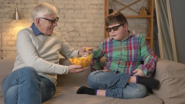 Молодой жадный мальчик берет миску чипсов у своего дедушки. Пожилой человек и молодой толстяк сидят на диване в 3D очках. Домашний комфорт, семейный уют, понятие уюта, разность — стоковое видео