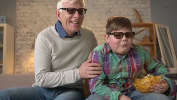 Grand-père et petit-fils sont assis sur le canapé et regardent un film en 3D dans des lunettes 3D, manger des chips, TV, spectacle. Confort à la maison, idylle familiale, concept de confort, différence de générations 60 ips — Video