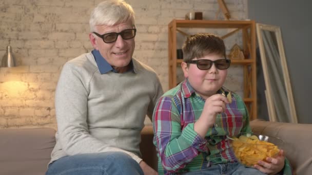 Grand-père et petit-fils sont assis sur le canapé et regardent un film en 3D dans des lunettes 3D, mangeant des chips, riant, souriant, comique, humour, télévision, spectacle. Confort à la maison, idylle familiale, concept de confort — Video