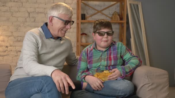 Nonno e nipote sono seduti sul divano e guardare un film 3D in bicchieri 3d, mangiare patatine fritte, TV, spettacolo. Comfort domestico, idillio familiare, concetto di comodità, differenza di generazioni. 60 fps — Video Stock