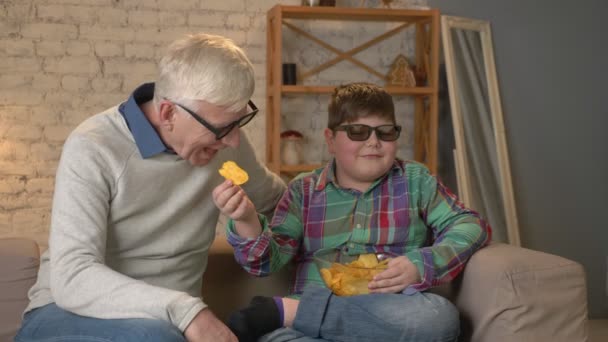Внук кормит дедушку чипсами из рук. Дедушка и внук сидят на диване и смотрят 3D-фильм в 3D-очках, едят чипсы, телевизор, показывают. Домашний комфорт, семейное благополучие — стоковое видео