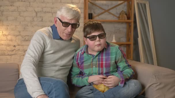 Grandfather and grandson are sitting on the couch and watching a 3D movie in 3d glasses, eating chips, moving, TV, show. Home comfort, family idyll, cosiness concept, difference of generations 60 fps — Stock Video