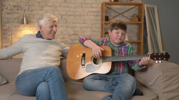 O neto toca para seu avô guitarra, música. Um homem idoso está sentado com um jovem gordo tocando guitarra. Conforto em casa, idílio familiar, conceito de aconchego, diferença de gerações 60 fps — Vídeo de Stock