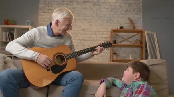 O avô toca para o neto na guitarra, na música. Um jovem gordo está sentado no refúgio com o avô. Ensina o neto a tocar guitarra. Conforto em casa, idílio familiar, conceito de conforto — Vídeo de Stock