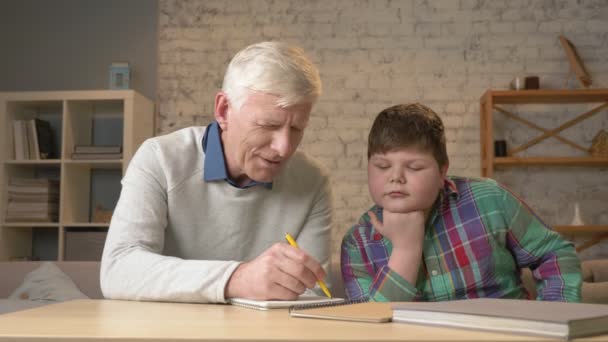 Il nonno aiuta un nipote con i compiti. L'uomo anziano aiuta un bambino grasso a fare i compiti. Comfort domestico, idillio familiare, concetto di comodità, differenza di generazioni, da vicino. 60 fps — Video Stock