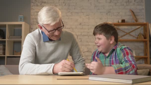 Opa hilft einem Enkel bei den Hausaufgaben. Ein älterer Mann hilft einem kleinen dicken Kind bei den Hausaufgaben. Wohnkomfort, Familienidylle, Gemütlichkeitskonzept, Generationenunterschied, hautnah. 60 fps — Stockvideo