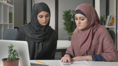 Hijabs iki güzel genç kız ofiste oturup tartışmak zamanlamaları, iş, diyalog, konuşma. 60 fps