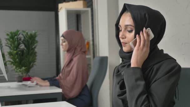 Jonge mooie verdrietig meisje in zwarte hijab zit in het kantoor en spreekt op de smartphone. Ongelukkig 60 fps — Stockvideo