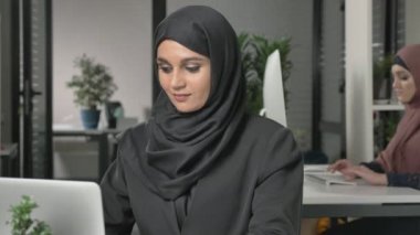 Siyah Tesettür office bilgisayar üzerinde çalışan genç güzel kız. Gülümseyen ve kameraya bakıyor. Arka planda kırmızı hijab kız. 60 fps kadar kapatın