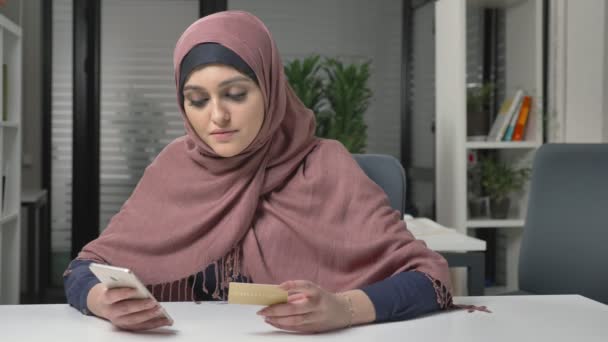 Joven chica hermosa en rosa hijab hace compras en Internet desde un teléfono inteligente. Tarjeta de crédito. 60 fps — Vídeos de Stock
