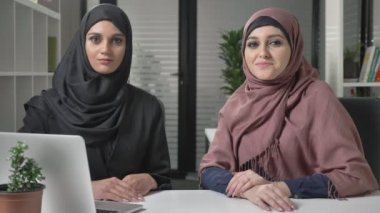 Hijab iki genç kız kameranın bir göz ile ofiste oturmak. Gülümseyerek. 60 fps