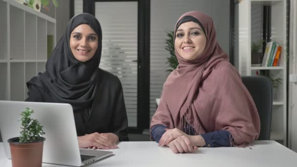 Two young girls in hijab sit in the office with a look at the camera. Smiling, positive 60 fps — Stock Video