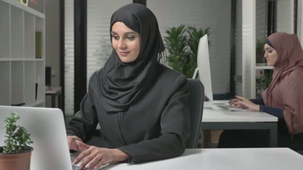 Ung vacker flicka i svart hijab maskinskrivning på laptop, titta på kameran och talar, talar. 60 fps — Stockvideo