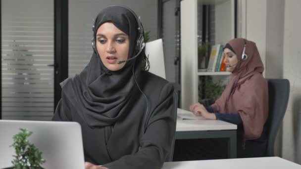 Mooie meisje in zwarte hijab met headset antwoorden een callcenter. Arabische vrouwen op kantoor. 60 fps — Stockvideo