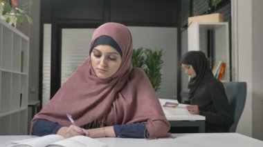 Genç güzel kız pembe bir hijab içinde bir defterde yazar öğrenir, çalışma. Arap kadınları ofiste. 60 fps