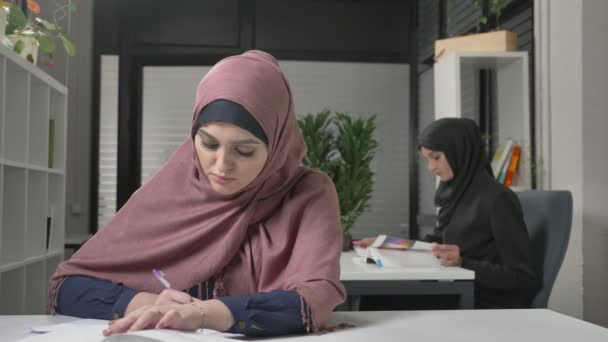 Mooie jonge meisje in een roze hijab schrijft in een notitieblok, leert, bestuderen. Arabische vrouwen op kantoor 60 fps — Stockvideo
