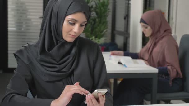 Menina bonita nova em hijab preto com fone de ouvido responde a um call center. Mulheres árabes no escritório. 60 fps — Vídeo de Stock