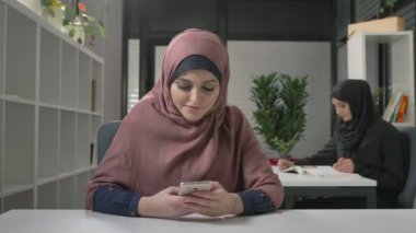 Kırmızı hijab güzel genç kızı ofiste oturur ve smartphone kullanır. Arap kadınları ofiste. 60 fps