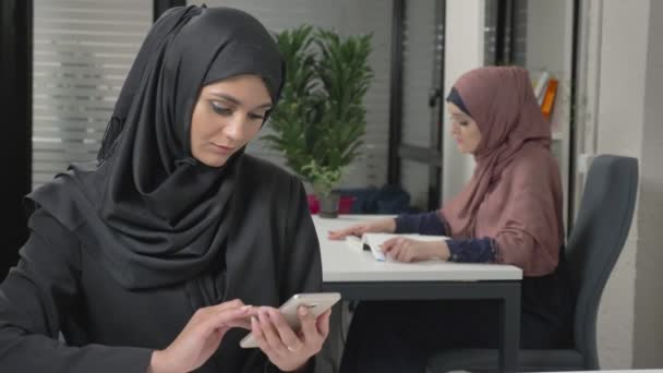 Joven chica hermosa en hijab negro con auriculares responde a un centro de llamadas. Mujeres árabes en la oficina. 60 fps — Vídeo de stock