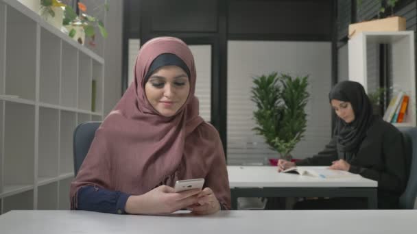 Belle jeune fille en hijab rouge se trouve dans le bureau et utilise un smartphone. Des femmes arabes dans le bureau. 60 ips — Video