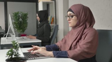 Genç güzel kız pembe hijab ve yazarak, bilgisayarda çalışarak gözlük. Kameraya bak. Arap kadınları ofiste. 60 fps