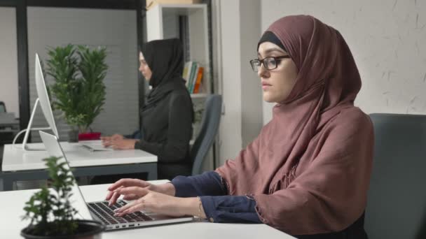 Ung vacker flicka i rosa hijab och glasögon maskinskrivning, arbetar på datorn. Titta på kameran. Arabiska kvinnor på kontoret. Flicka i svart hijab i bakgrunden. 60 fps — Stockvideo