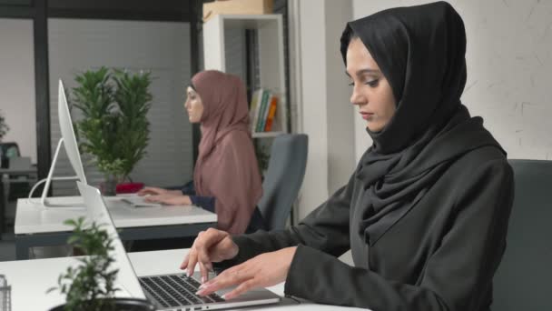 Junges schönes Mädchen im schwarzen Hijab, das im Büro am Laptop arbeitet, massiert seinen Whisky, Migräne, Kopfschmerzen. Frau in rosa Hidschab im Hintergrund. 60 fps — Stockvideo