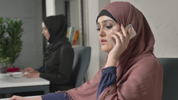 Joven chica hermosa en hijab rosa se sienta en la oficina y habla en el teléfono inteligente, riendo. 60 fps — Vídeo de stock