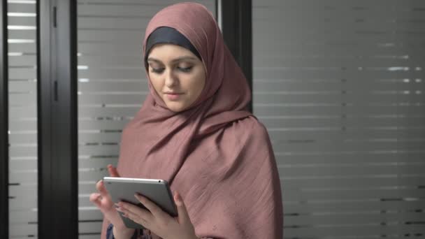 Een mooi meisje in roze hijab maakt gebruik van een tablet, kijkt naar de foto's. Arabische vrouwen op kantoor. 60 fps — Stockvideo