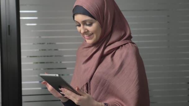 Uma jovem garota bonita em hijab rosa usa um tablet, olha para fotos, sorrindo. Mulheres árabes no escritório. 60 fps — Vídeo de Stock