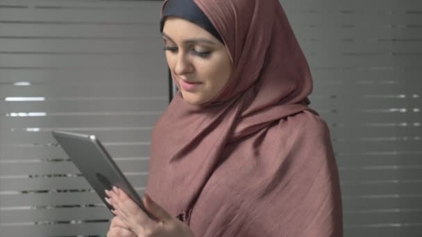 Ein junges schönes Mädchen im rosafarbenen Hijab bedient sich eines Tablets, spricht in einem Videochat und grüßt. 60 fps — Stockvideo