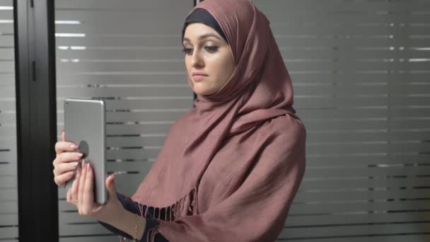 Ein junges schönes Mädchen im rosafarbenen Hijab bedient sich eines Tablets, spricht in einem Videochat und grüßt. 60 fps — Stockvideo