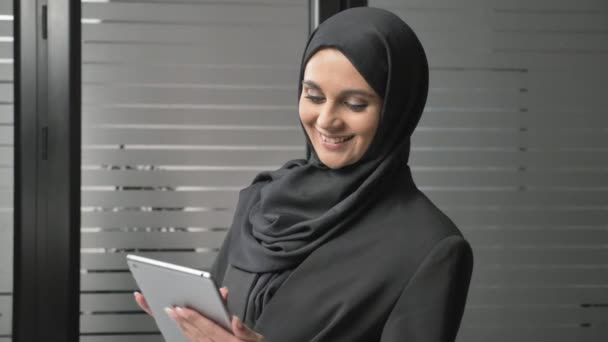 Seorang gadis cantik mengenakan jilbab hitam menggunakan tablet, melihat foto, tersenyum. Wanita Arab di kantor. 60 fps — Stok Video