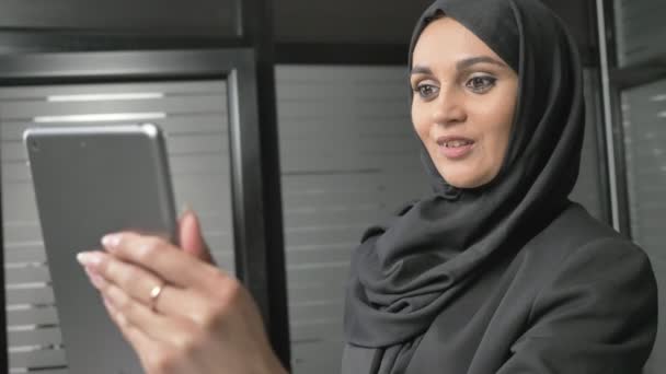 A young beautiful girl in black hijab uses a tablet, speaks in a video chat, greeting. 60 fps — Stock Video