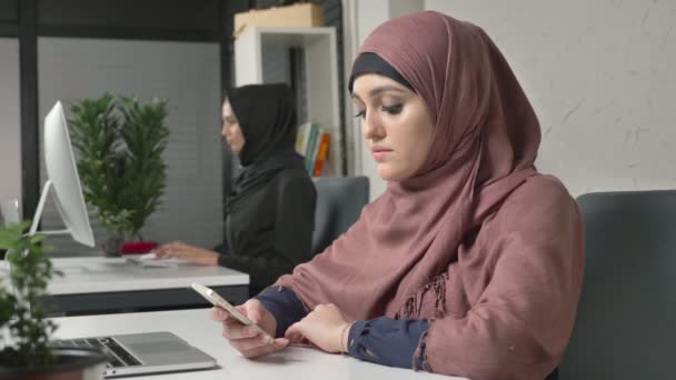 Krásná mladá dívka v růžové hidžáb sedí v kanceláři a používá smartphone. Dívka v černém hidžáb na pozadí. Arabské ženy v kanceláři. 60 snímků za sekundu — Stock video