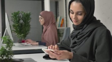 Siyah Tesettür genç güzel kızı ofiste oturur ve smartphone kullanır. Arka planda siyah hijab kız. Arap kadınları ofiste. 60 fps