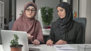 Hijabs iki güzel genç kız ofiste oturup tartışmak zamanlamaları, iş, diyalog, konuşma. 60 fps