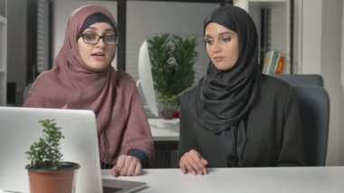 Hijabs iki güzel genç kız ofiste oturup tartışmak zamanlamaları, iş, diyalog, konuşma. 60 fps