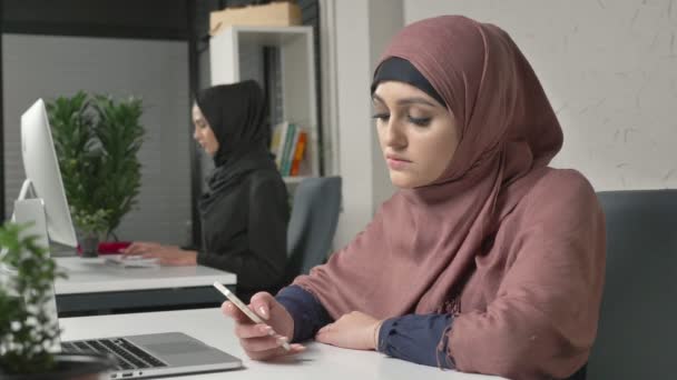 Mooie meisje in roze hijab zit in office en maakt gebruik van de smartphone. Meisje in zwarte hijab in de achtergrond. Arabische vrouwen op kantoor. 60 fps — Stockvideo