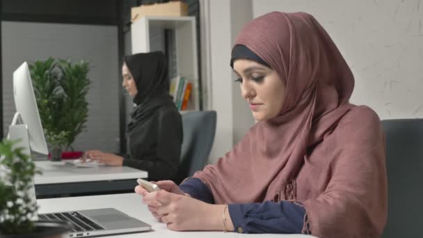 Όμορφη κοπέλα με ροζ χιτζάμπ κάθεται στο γραφείο και χρησιμοποιεί smartphone. Κορίτσι στο μαύρο μαντίλα στο παρασκήνιο. Αραβικά γυναίκες στο γραφείο. 60 fps — Αρχείο Βίντεο