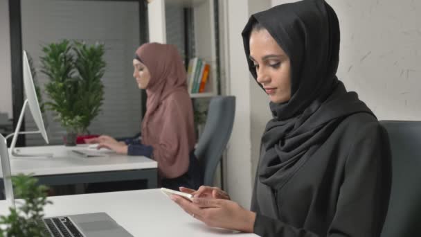 Junges schönes Mädchen im schwarzen Hijab sitzt im Büro und benutzt Smartphone. Mädchen im schwarzen Hijab im Hintergrund. Araberinnen im Büro. 60 fps — Stockvideo