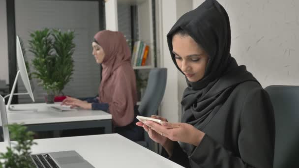 Joven chica hermosa en negro hijab se sienta en la oficina y utiliza el teléfono inteligente. Chica en hiyab negro en el fondo. Mujeres árabes en la oficina. 60 fps — Vídeo de stock