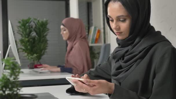 Jeune belle fille en hijab noir se trouve dans le bureau et utilise un smartphone. Fille en hijab noir en arrière-plan. Des femmes arabes dans le bureau. 60 ips — Video
