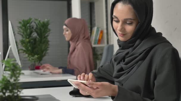 Gadis cantik mengenakan jilbab hitam duduk di kantor dan menggunakan smartphone. Gadis berhijab hitam di latar belakang. Wanita Arab di kantor. 60 fps — Stok Video