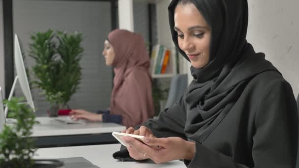 Όμορφη κοπέλα με μαύρο χιτζάμπ κάθεται στο γραφείο και χρησιμοποιεί smartphone. Κορίτσι στο μαύρο μαντίλα στο παρασκήνιο. Αραβικά γυναίκες στο γραφείο. 60 fps — Αρχείο Βίντεο