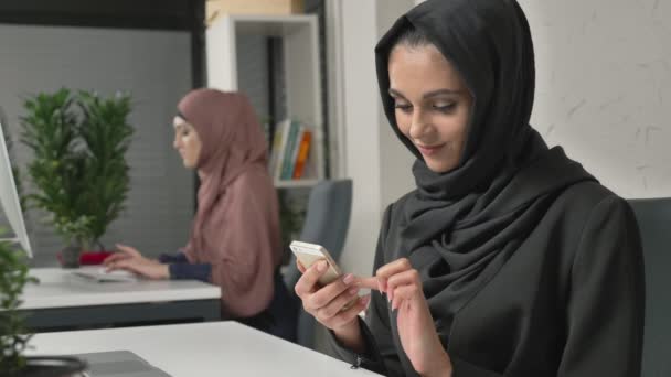 Jeune belle fille en hijab noir se trouve dans le bureau et utilise un smartphone. Fille en hijab noir en arrière-plan. Des femmes arabes dans le bureau. 60 ips — Video