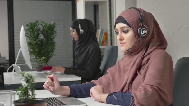 Seorang gadis cantik mengenakan jilbab merah muda sedang berbicara di headset, menjawab panggilan di pusat panggilan. Wanita Arab di kantor. 60 fps — Stok Video