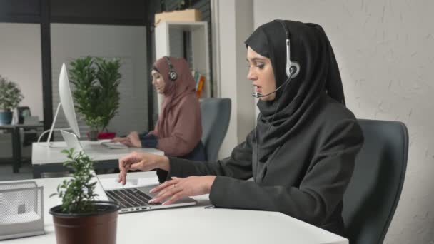 Una joven hermosa en un hiyab negro está hablando en los auriculares, respondiendo a las llamadas en el centro de llamadas. Mujeres árabes en la oficina. 60 fps — Vídeo de stock