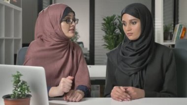 Hijabs iki güzel genç kız ofiste oturup tartışmak zamanlamaları, iş, diyalog, konuşma. 60 fps