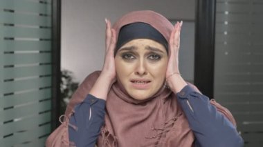 Genç üzücü Arap kız pembe hijab elleriyle kulaklarını kapatır. Kameraya, görünüyor portre. 60 fps